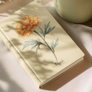 Könnte beinhalten: Ein cremefarbenes Notizbuch mit floralem Design. Das Cover zeigt eine detaillierte Illustration einer gelb-orangenen Pfingstrose mit grünen Blättern und dem Namen "Elijah" in Schreibschrift. Das Notizbuch liegt auf einer weißen Oberfläche.