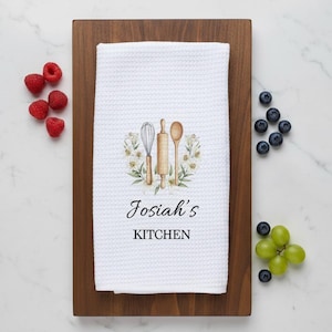 May include: White waffle-weave kitchen towel with a watercolor design of a whisk, rolling pin, and spoon surrounded by flowers. The text "Josiah's KITCHEN" is printed below the design. The towel is on a wooden cutting board, with raspberries and blueberries.