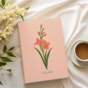 May include: A pale pink notebook with a floral design of orange gladioli and green leaves. The name "LEILANI" is printed at the bottom. The notebook rests on a white satin surface, with a cup of coffee and flowers in the background.