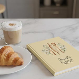May include: A light yellow recipe book with the text "Josiah's Favourite Recipes" and illustrations of kitchen tools. A croissant sits on a white plate next to a glass mug of coffee.