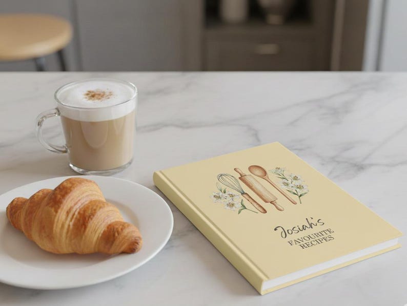 May include: A light yellow recipe book with the text "Josiah's Favourite Recipes" and illustrations of kitchen tools. A croissant sits on a white plate next to a glass mug of coffee.