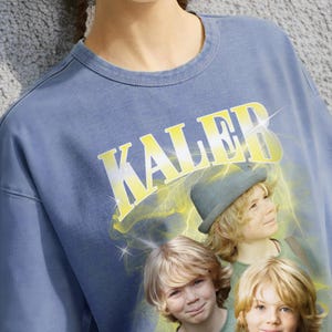 May include: Blue crew neck sweatshirt with the word "Kaleb" in yellow block letters. The graphic features three children with blonde hair and a hat, with a yellow lightning bolt design behind them. The sweatshirt is a solid blue color.