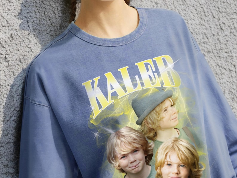 May include: Blue crew neck sweatshirt with the word "Kaleb" in yellow block letters. The graphic features three children with blonde hair and a hat, with a yellow lightning bolt design behind them. The sweatshirt is a solid blue color.