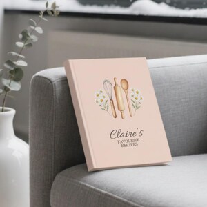 May include: A light pink recipe book titled "Claire's Favourite Recipes" with a watercolor illustration of kitchen utensils. The book is propped on a gray upholstered armchair next to a white vase with eucalyptus branches.