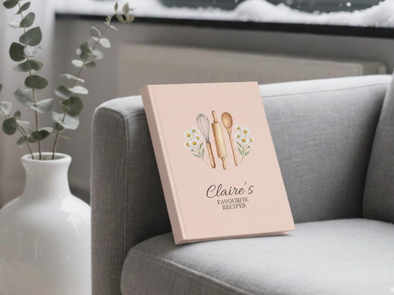 May include: A light pink recipe book titled "Claire's Favourite Recipes" with a watercolor illustration of kitchen utensils. The book is propped on a gray upholstered armchair next to a white vase with eucalyptus branches.