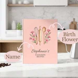May include: A pink recipe book with the text "Stephanie's Favourite Recipes" and a floral design. The cover features a whisk, rolling pin, and spoon. The book is on a white countertop with a black mug and coffee beans.