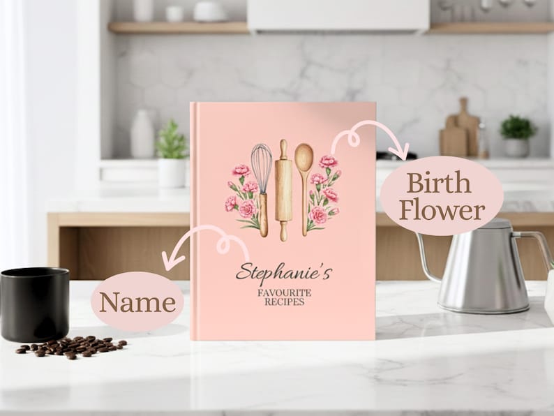 May include: A pink recipe book with the text "Stephanie's Favourite Recipes" and a floral design. The cover features a whisk, rolling pin, and spoon. The book is on a white countertop with a black mug and coffee beans.