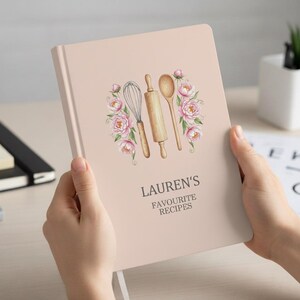 Può includere: Un quaderno di ricette rosa chiaro con il testo "LAUREN'S FAVOURITE RECIPES". La copertina presenta un'illustrazione ad acquerello di una frusta, un mattarello e un cucchiaio, circondati da fiori rosa. Il quaderno è tenuto da una persona.
