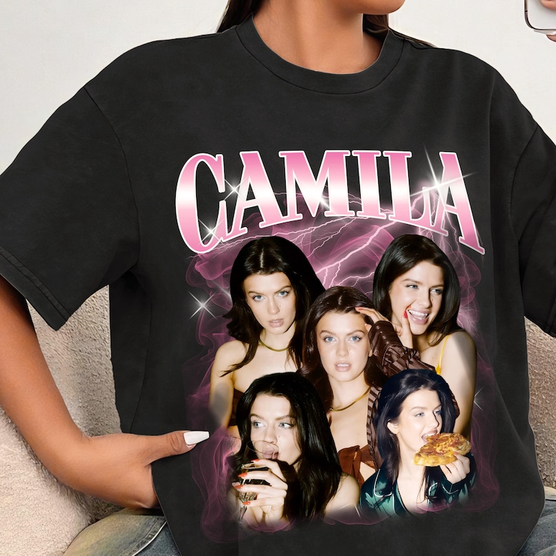 May include: Black t-shirt featuring a graphic of five women with the name "CAMILA" in pink, outlined in white. The graphic has a pink lightning bolt design in the background.