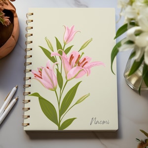 May include: A cream-coloured, spiral-bound notebook with a pink lily floral design. The name "Naomi" is printed in a script font. Two white pens with gold accents are next to the notebook.