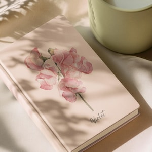 Peut inclure: Un carnet rose avec un motif floral à l'aquarelle de fleurs roses. Le mot "Violet" est écrit en cursive sur la couverture. Une tasse en céramique vert clair est en arrière-plan.