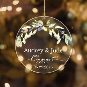 Könnte beinhalten: Ein klarer, runder Christbaumschmuck mit einem goldenen Aufhänger. Er zeigt die Namen "Audrey & Jude", das Wort "Engaged" und das Datum "06.29.2025". Die Oberseite ist mit einem Aquarelldesign aus Olivenzweigen und Oliven verziert.