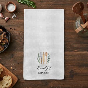 May include: White waffle-weave kitchen towel featuring a watercolour design of a whisk, rolling pin, and spoon, surrounded by greenery. The text "Emily's KITCHEN" is printed below the design. The towel is displayed on a wooden surface.