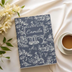 May include: A blue notebook with a white toile pattern and the name "Camila" printed on the front. The notebook is lying on a white surface with white flowers and a cup of coffee.