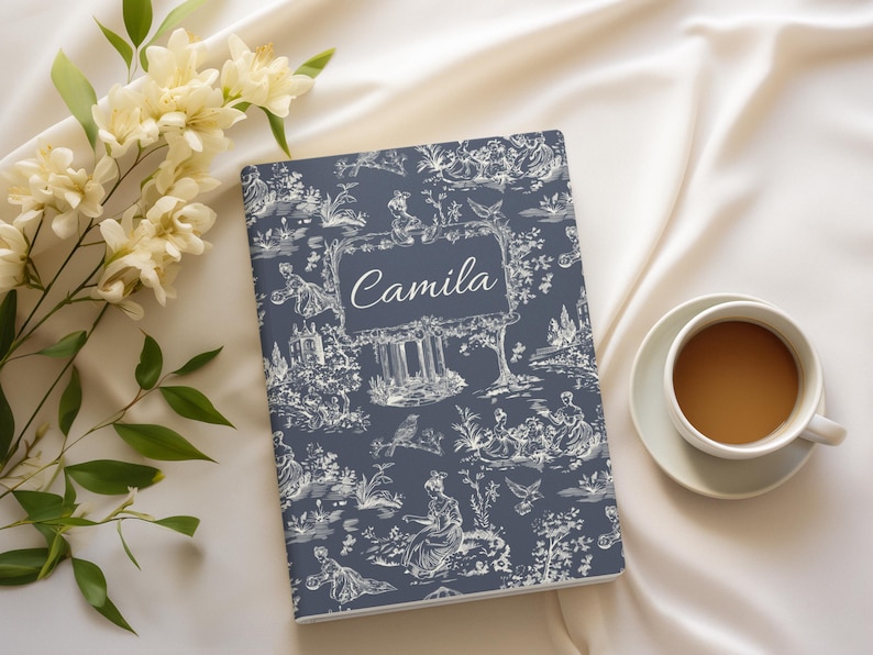 May include: A blue notebook with a white toile pattern and the name "Camila" printed on the front. The notebook is lying on a white surface with white flowers and a cup of coffee.
