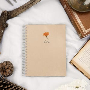Peut inclure: Un carnet à spirales beige avec une fleur orange et le nom "Luna" écrit en cursive. Le carnet est entouré d'un livre, d'une pomme de pin et d'une branche.