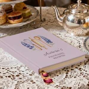May include: A lavender-colored recipe book titled "Aurora's Favourite Recipes" with a watercolor illustration of kitchen tools. The book is open with a bookmark and dried rose petals. A silver teapot and tiered serving tray are in the background.
