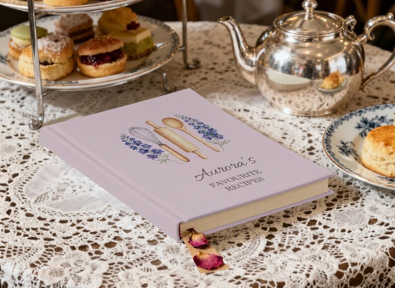May include: A lavender-colored recipe book titled "Aurora's Favourite Recipes" with a watercolor illustration of kitchen tools. The book is open with a bookmark and dried rose petals. A silver teapot and tiered serving tray are in the background.