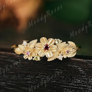 以下が含まれることがあります： 中央に赤い宝石をあしらった花、周囲にピンクと白の宝石をあしらった小さな花が咲く、金色のフローラルリング。 リングには葉のディテールがあり、濃い木製の表面にセットされています。
