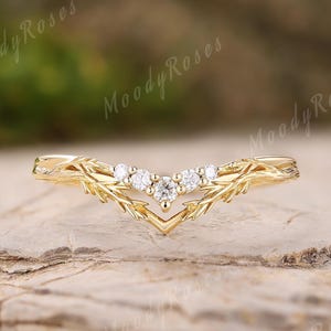 Könnte beinhalten: Ein goldfarbener Ring mit einem blattartigen Design und einem V-förmigen Band. Der Ring hat fünf kleine, klare Edelsteine in der Mitte. Der Ring wird auf einer strukturierten, braunen Oberfläche präsentiert.