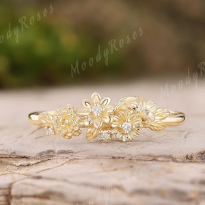 May include: A gold-colored ring featuring a floral design with small, sparkling gemstones. The ring's band is thin, and the floral arrangement is clustered at the top. The ring is set against a natural, textured background.