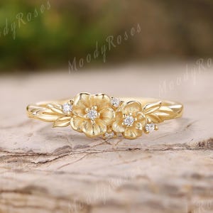 May include: A gold ring featuring a floral design with two prominent flowers and several small diamonds. The ring has a delicate, nature-inspired aesthetic, with leaves and additional diamond accents. The ring is set on a textured, light brown surface.