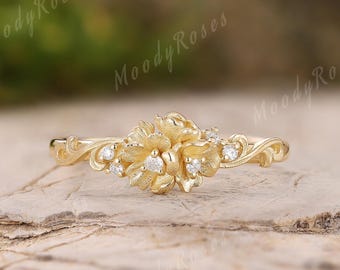 May include: A gold-colored ring with a floral design. The ring features a cluster of small, round gemstones at the center of the flower and along the band. The ring is set against a neutral background.