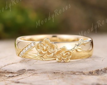 May include: A gold-colored ring with a floral design. The ring features two sculpted flowers and a row of small, clear stones. The band is smooth and polished. The ring is set against a natural, textured background.