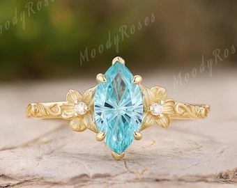 May include: A gold engagement ring with a marquise-cut, light blue gemstone. The ring features floral accents with small white gemstones on each side of the center stone. The band has an engraved design.