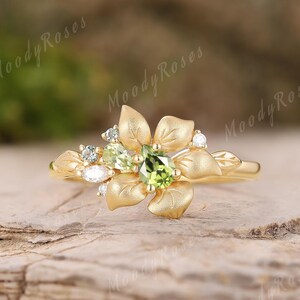 Op de afbeelding: Een goudkleurige ring met een bloemmotief. De ring heeft een centrale peridot edelsteen, omringd door kleinere witte en groene edelstenen. De band en bloemblaadjes hebben een getextureerde afwerking. De ring ligt op een houten oppervlak.