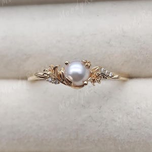May include: A gold ring featuring a central white pearl flanked by leaf-shaped metal accents and small clear stones. The band has a polished finish and the text Moody Roses is visible in the background. The design incorporates organic, botanical-inspired metalwork.