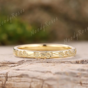 Puede incluir: Un anillo dorado con un patrón floral. El anillo está colocado sobre una superficie de piedra natural. El diseño del anillo presenta grabados detallados de flores y hojas, creando una estética delicada y elegante. El anillo es un símbolo de compromiso.
