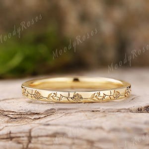 Może przedstawiać: Złoty pierścionek z grawerowanym wzorem kwiatowym wokół obrączki. Pierścionek leży na naturalnej drewnianej powierzchni. Słowa "Moody Roses" są słabo widoczne w tle.