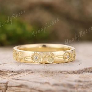 May include: A gold band ring with a floral design. The ring features engraved sunflowers and small diamond accents. The ring is set on a natural stone surface. The text "Moody Roses" is visible in the background.