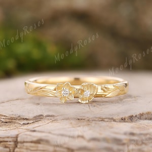 Op de afbeelding: Een gouden ring met bloemmotieven en twee bloemvormige accenten, elk met een kleine, heldere edelsteen. De ring wordt getoond op een natuurlijke, getextureerde achtergrond. De woorden "Moody Roses" zijn zichtbaar op de achtergrond.