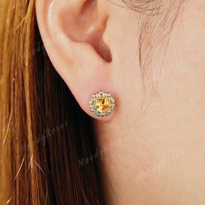 May include: A close-up of a stud earring featuring a central, round, yellow gemstone surrounded by smaller, clear stones. The earring has a gold-colored setting and is set against the backdrop of a person's ear.