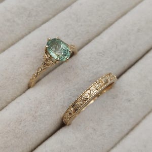 May include: Two gold rings. One ring features an oval, light green gemstone. The other ring is a gold band with an engraved floral pattern. Both rings are displayed on a light gray surface.