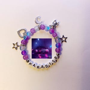 May include: A purple and blue beaded bracelet with a charm that says "PARADISE" and a small image of a car. The bracelet also has a crescent moon charm, a star charm, and a heart charm.