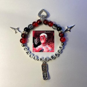 Puede incluir: Un brazalete de cuentas rojas y negras con un dije plateado de un teléfono plegable. El brazalete tiene cuentas blancas con las palabras "CALL ME BACK" escritas. El brazalete también tiene un dije de corazón plateado y dos dijes de estrella plateados. El brazalete está centrado en un fondo blanco y tiene una pequeña portada de álbum en el fondo.