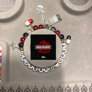 Puede incluir: Un brazalete de cuentas blancas y negras con las palabras "Drugs and Money" escritas. El brazalete también tiene cuentas rojas y negras, un dije de guitarra plateado, un dije de billete de 100 euros plateado y un dije de estrella plateado. El brazalete está sobre una superficie blanca con una regla en el fondo.