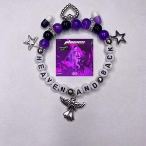 Puede incluir: Una pulsera de cuentas con las palabras "HEAVEN AND BACK" escritas con cuentas de letras blancas. La pulsera presenta cuentas moradas y negras, dijes de estrella y un dije de ángel plateado. Una pequeña portada de álbum está en el centro.