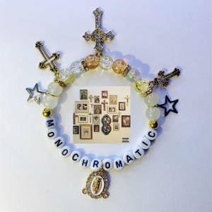 May include: A beaded bracelet with the word "MONOCHROMATIC" spelled out in white letter beads. The bracelet features gold-colored crosses, stars, and a pendant. The center of the bracelet has a small image of framed pictures.