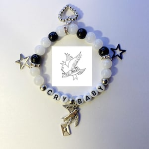 May include: White and black beaded bracelet with a silver charm of a bird holding a letter. The bracelet has the words "CRY BABY" spelled out on it and a small silver heart charm. The bracelet also has a black star charm and a black and white image of a dove with the words "all merch" written on it.
