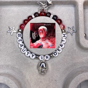 Puede incluir: Un brazalete de cuentas con cuentas blancas que deletrean "BEAUTY IN DEATH" y cuentas rojas y negras. El brazalete tiene un dije de calavera plateado y un dije plateado con un diseño de mariposa. También tiene dos dijes de estrella plateados. El brazalete está centrado en una portada de álbum roja y negra con el texto "BEAUTY BEHIND THE MADNESS" en ella.