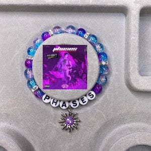 Puede incluir: Un brazalete de cuentas con un esquema de color morado, azul y blanco. El brazalete tiene un dije con la palabra "PHASES" y un sol con una gema morada en el centro.