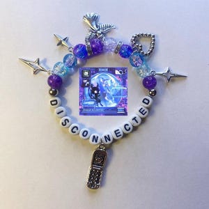 May include: A white beaded bracelet with the word "DISCONNECTED" spelled out in black letters. The bracelet has several charms, including a silver star, a silver heart, a silver phone, and a silver angel wing. The bracelet also has a blue and purple card with a black and white image of a creature.