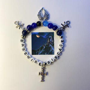 May include: A white beaded bracelet with the words "Barbwire Ribcage" spelled out. The bracelet has blue and black beads, a silver cross charm, and a silver angel wing charm.