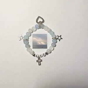 May include: A white and blue beaded bracelet with a silver cross charm and the name "Lillie" spelled out in white beads. The bracelet also has two silver star charms.