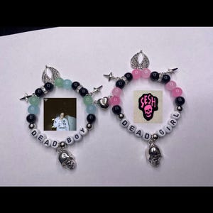 Puede incluir: Dos pulseras de cuentas con dijes de calavera. Una pulsera tiene cuentas negras, rosadas y blancas con el texto "DEAD GIRL" y un dije que dice "SESH". La otra pulsera tiene cuentas negras, verdes y blancas con el texto "DEAD BOY".