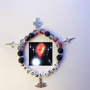 May include: A beaded bracelet with black and red beads, a silver cross charm, a silver star charm, and a charm with the word "RED LIGHT" spelled out in white letters.